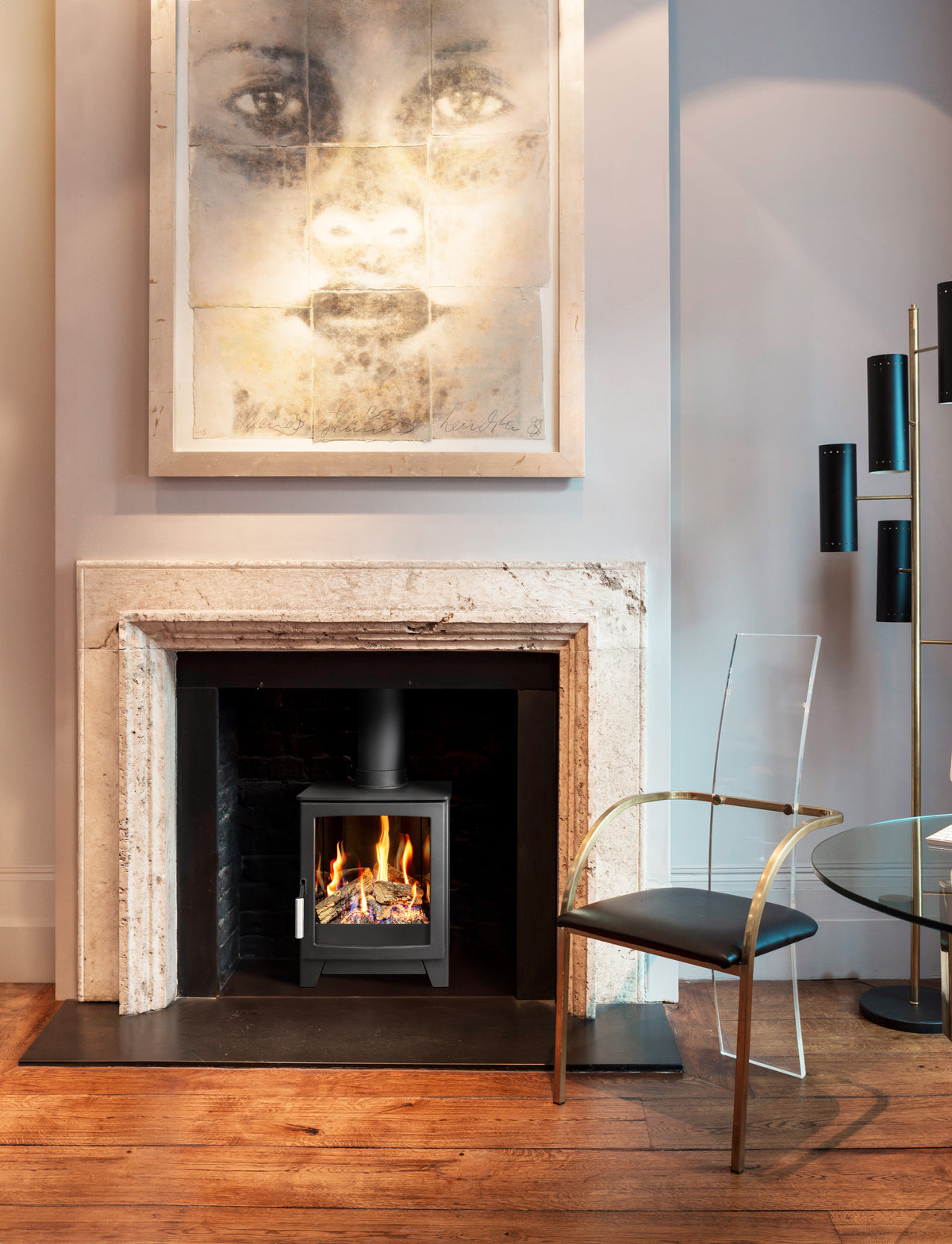 A modern black gas stove fireplace insert with visible flames, set within a light stone fireplace surround in a styled interior. A large framed portrait hangs above the fireplace, and a gold-framed chair with a black seat is positioned to the right.