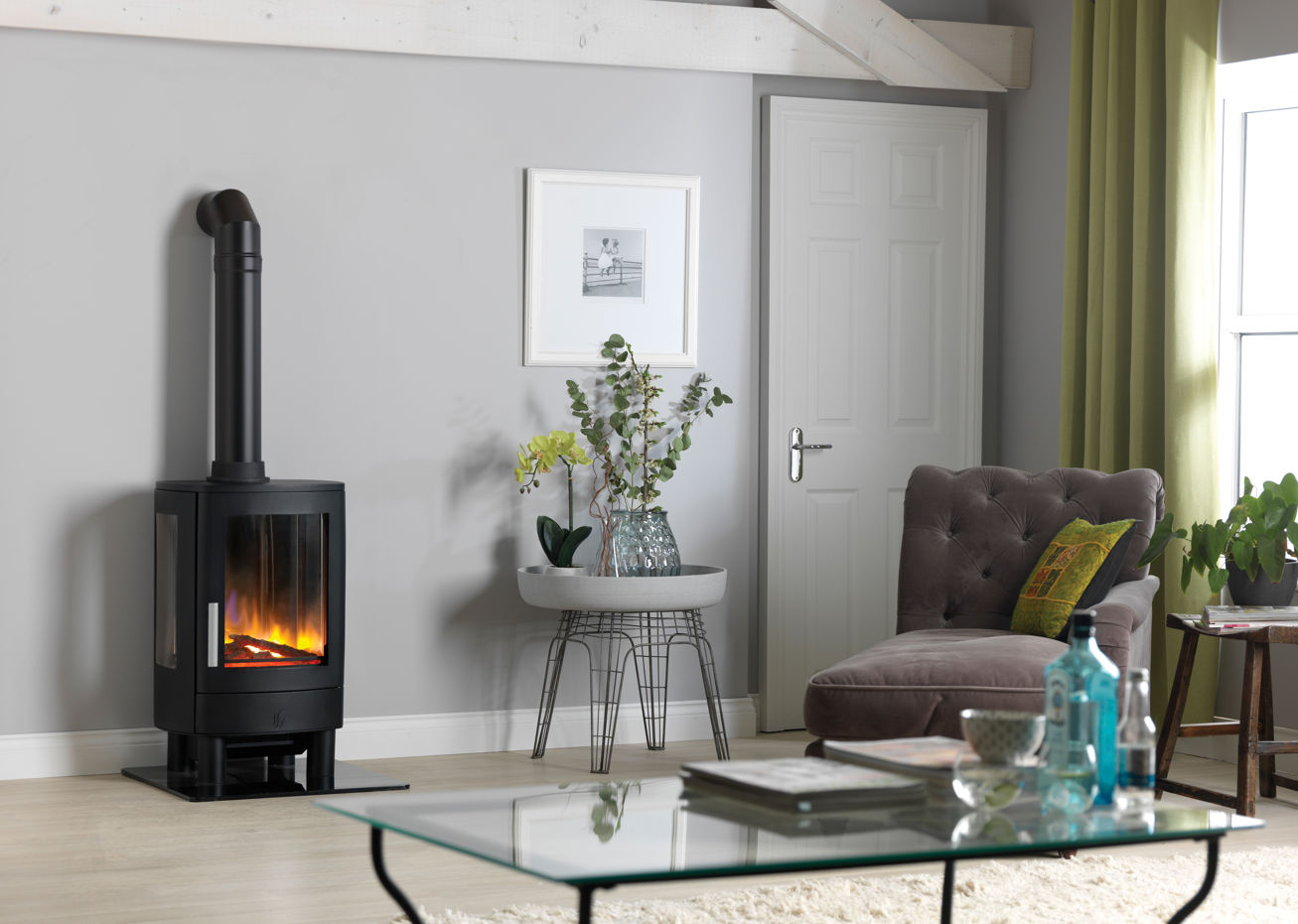 A black wood-burning stove with a visible fire, set in a modern living room next to a gray armchair, glass coffee table, and green curtains.