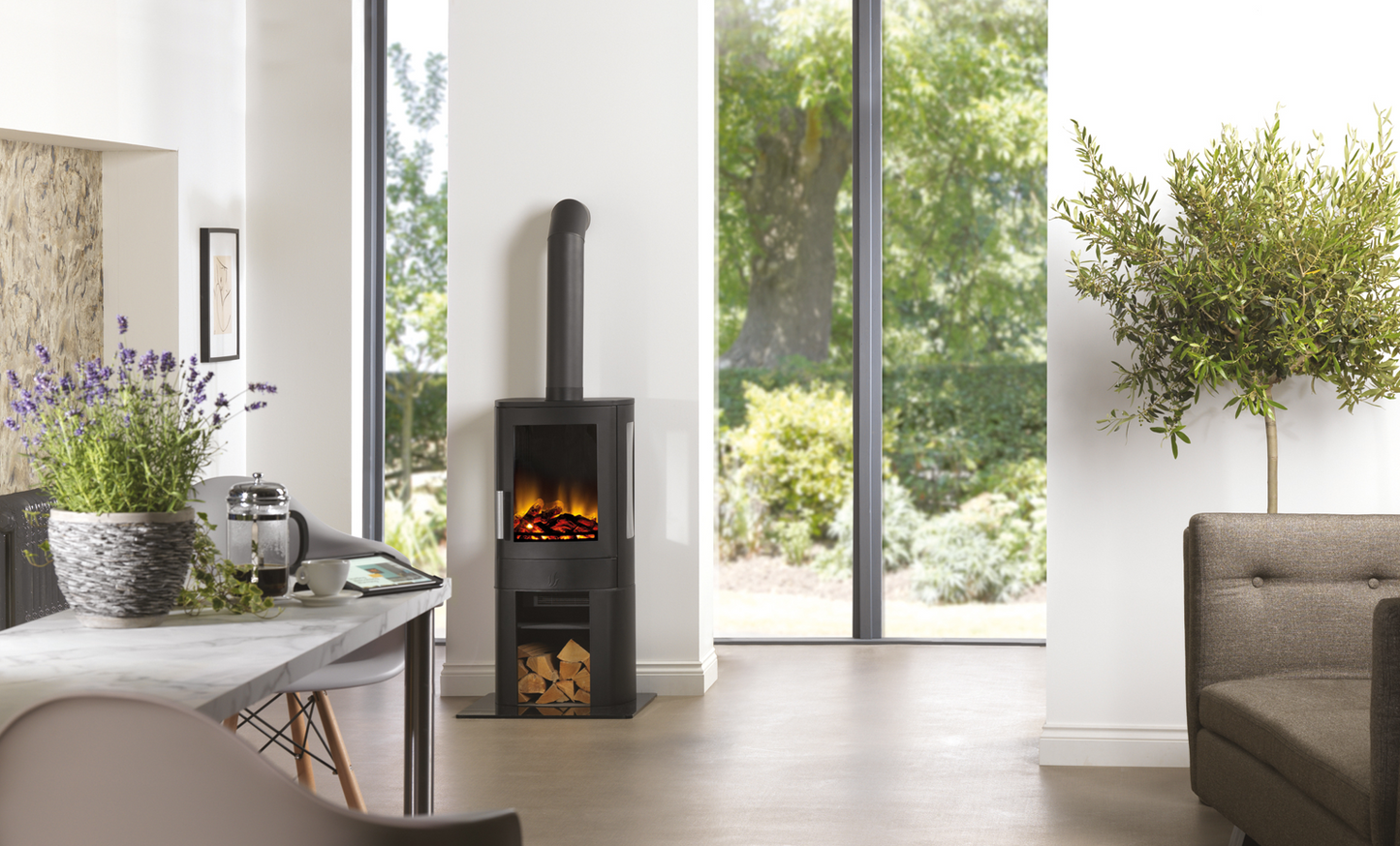 A modern black wood-burning stove with a visible fire, set in a bright, contemporary living room with large windows looking out to greenery.