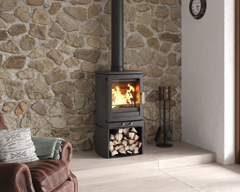 A black wood-burning stove with an open fire visible in the glass front, set against a stone wall in a room with a light wood floor. A brown leather armchair with patterned pillows is partially visible on the left, and fireplace tools stand on the right.