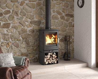 A black wood-burning stove with an open fire visible in the glass front, set against a stone wall in a room with a light wood floor. A brown leather armchair with patterned pillows is partially visible on the left, and fireplace tools stand on the right.