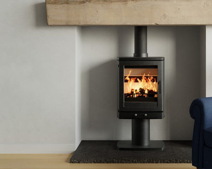 A black wood-burning stove with a visible fire inside, set against a light gray wall beneath a rustic wooden mantel. A dark floor mat sits below the stove, and part of a blue upholstered chair is visible on the right.
