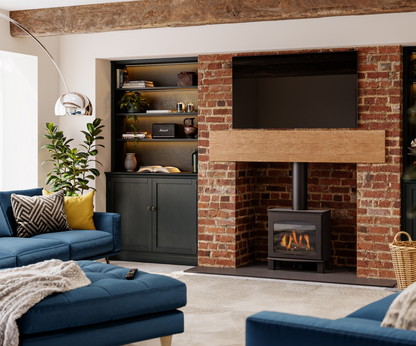 A modern living room featuring a black wood-burning stove insert set into a red brick fireplace with a light wood mantel. The room includes a blue sofa, a matching ottoman, a built-in dark cabinet with shelves, and a large floor lamp.