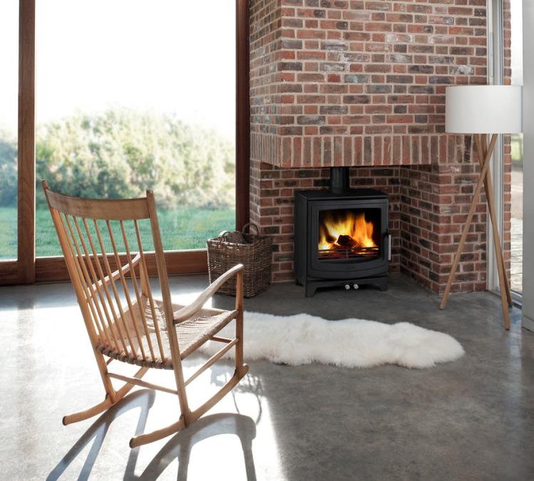 AGA Ellesmere Multi-Fuel Stove. Wooden rocking chair in front of a lit fireplace with a brick wall and large windows.