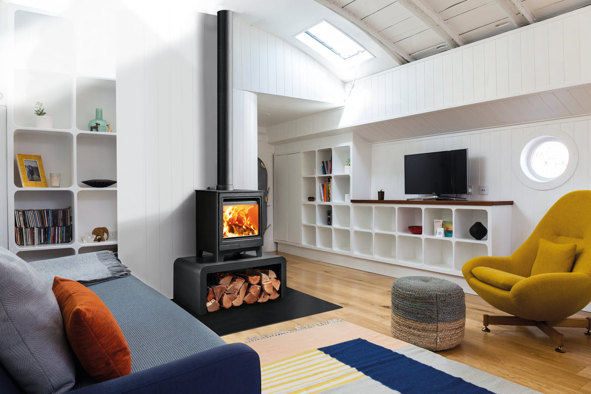 A modern wood-burning stove with a black chimney stack and fire visible in the glass front, installed in a loft-style living room with white shelving, a blue sofa, a yellow armchair, and a striped rug.