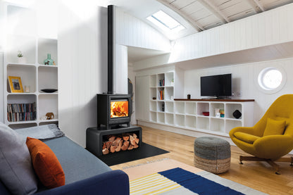 A modern wood-burning stove with a black chimney stack and fire visible in the glass front, installed in a loft-style living room with white shelving, a blue sofa, a yellow armchair, and a striped rug.