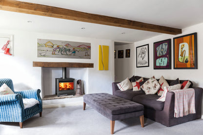 A black wood-burning stove with a glass front door sitting on a bench with logs beneath showing a bright fire and logs, in a lifestyle shot in a cosy lounge.