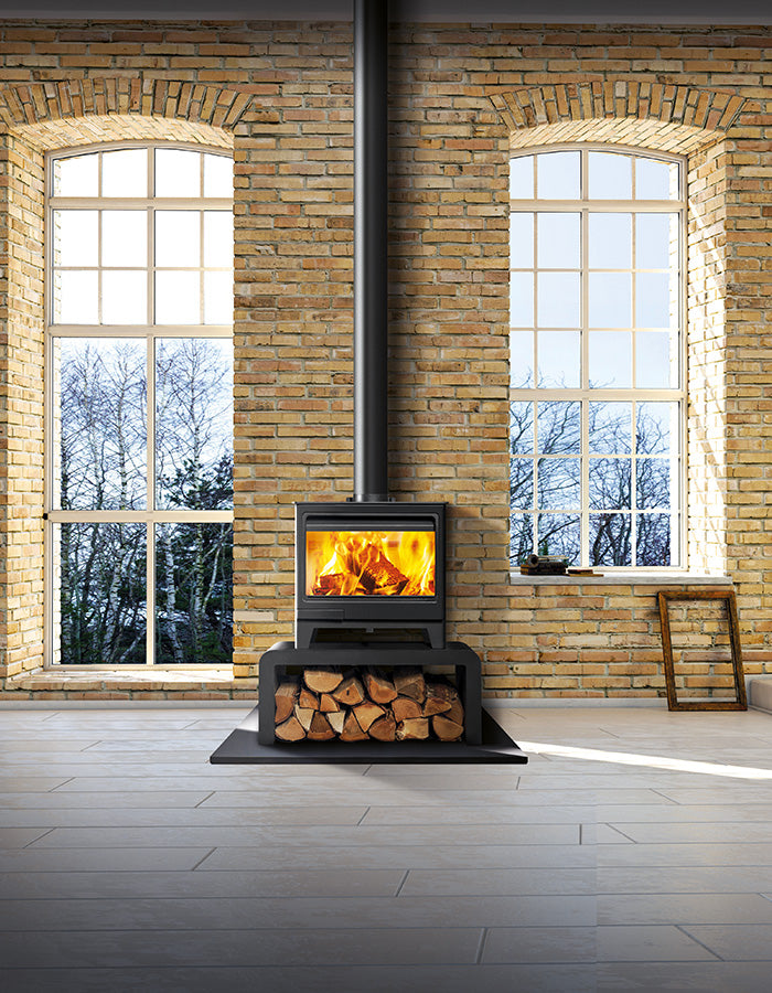 A black wood-burning stove with a glass front door sitting on a bench with logs beneath showing a bright fire and logs, in a lifestyle shot with windows and trees.