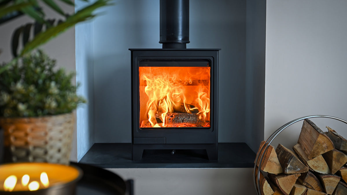 A black wood-burning stove with a glowing orange fire inside, set against a dark gray wall with a chimney pipe extending upward. A bowl of lit candles sits in the blurred foreground on the left, and a basket of firewood is visible on the right.