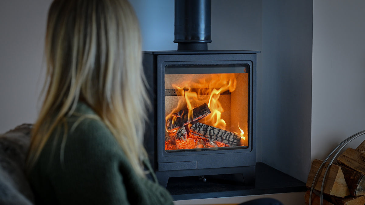 A woman in a cozy green sweater sits in the foreground, viewed from behind, looking at a burning wood-burning stove with bright orange flames and glowing embers. The stove has a black metal body and a tall black chimney pipe, set against a dark gray wall with stacked firewood visible on the right.