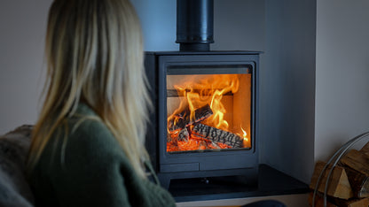 A woman in a cozy green sweater sits in the foreground, viewed from behind, looking at a burning wood-burning stove with bright orange flames and glowing embers. The stove has a black metal body and a tall black chimney pipe, set against a dark gray wall with stacked firewood visible on the right.