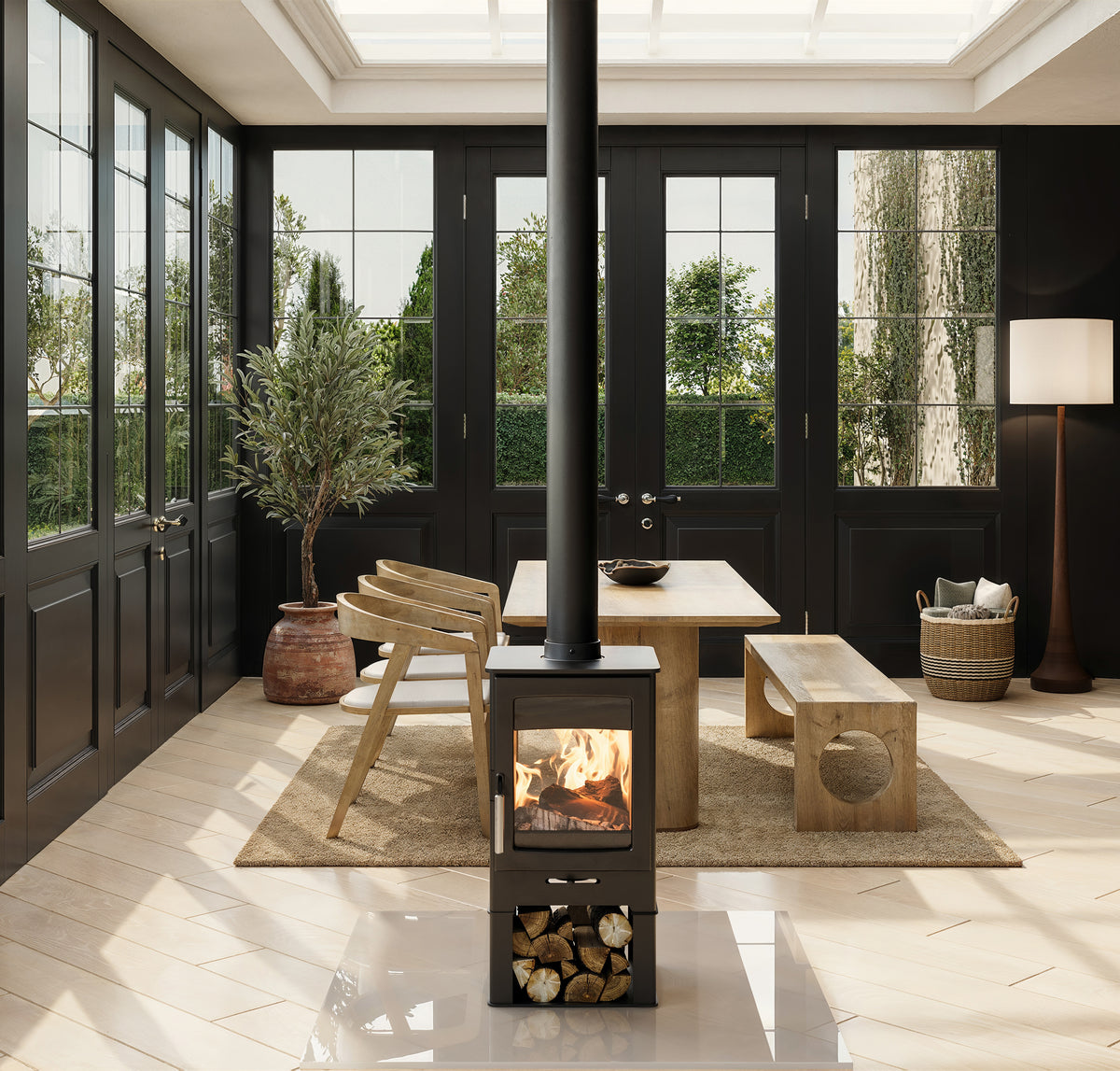 A modern black wood-burning stove with a visible fire, styled in a bright, sunlit sunroom with black-framed glass doors, a wooden dining table, chairs, and a long bench.