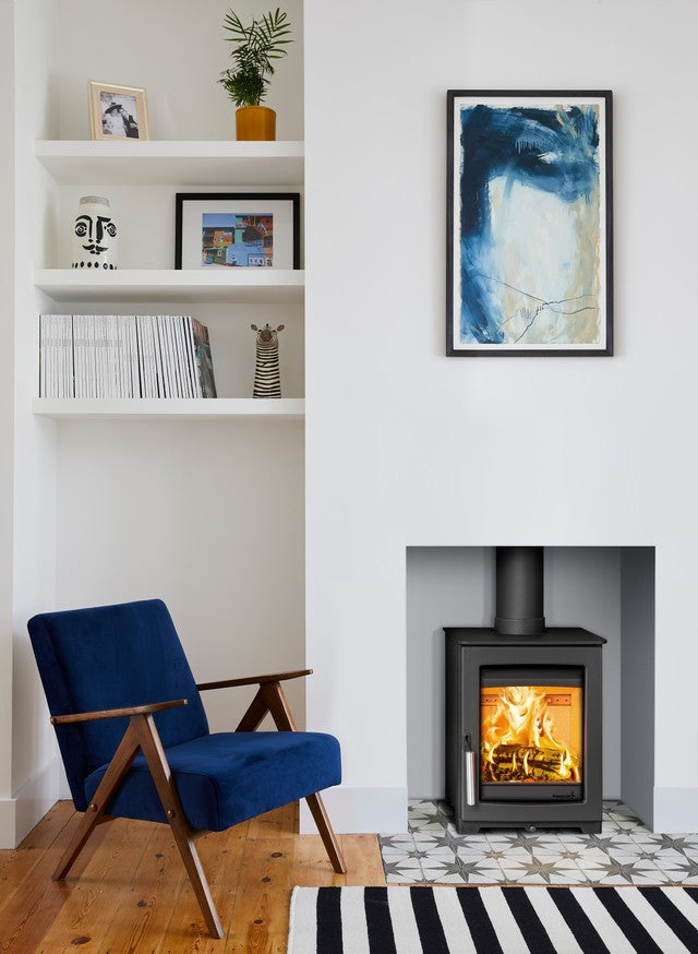 A modern living room corner featuring a black wood-burning stove with a glowing fire, a blue velvet armchair, white built-in shelves, and a framed abstract blue painting on a white wall.