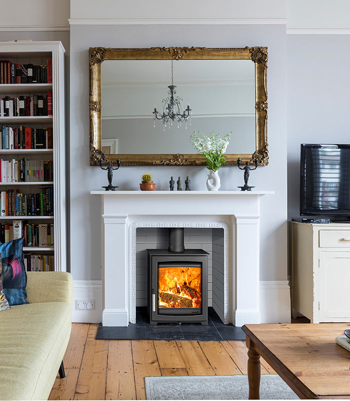 A large ornate gold-framed rectangular mirror hangs on a white wall above a white fireplace mantel with a wood-burning stove. The room includes a bookshelf, sofa, television, and wooden floor.