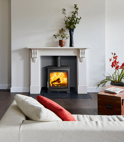 A modern black wood-burning stove with a glowing fire, set within a white fireplace mantel in a styled living room with a sofa, pillows, and decorative plants.