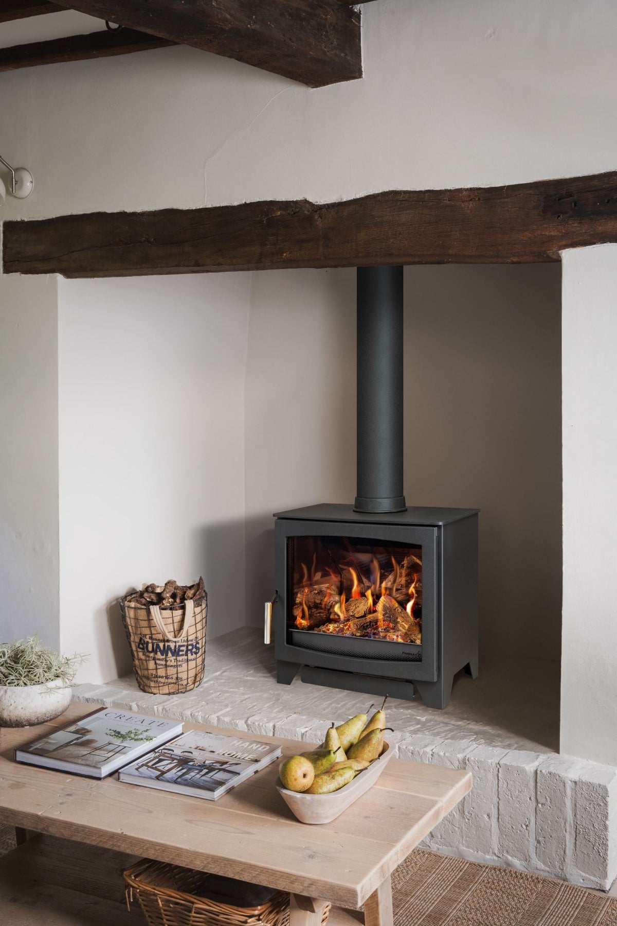 A black gas stove with a visible fire, set in a rustic room with a stone hearth, a wooden table with magazines and pears, and a basket of firewood beneath a dark wooden beam.