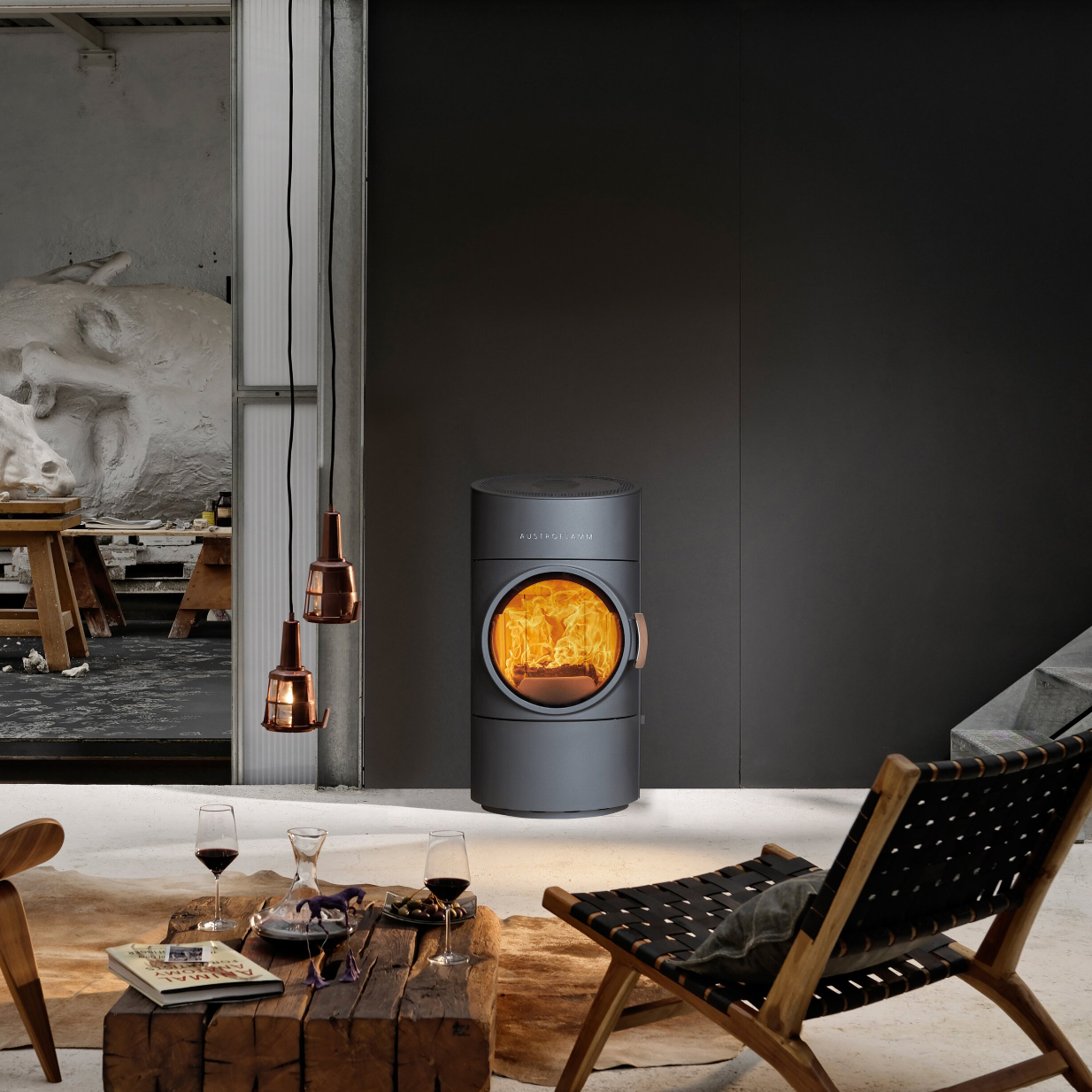 A modern dark gray Pelton-branded wood-burning stove with a glowing fire visible through its circular glass door, set in a stylish contemporary interior with a wooden chair, table, and hanging copper pendant lights.