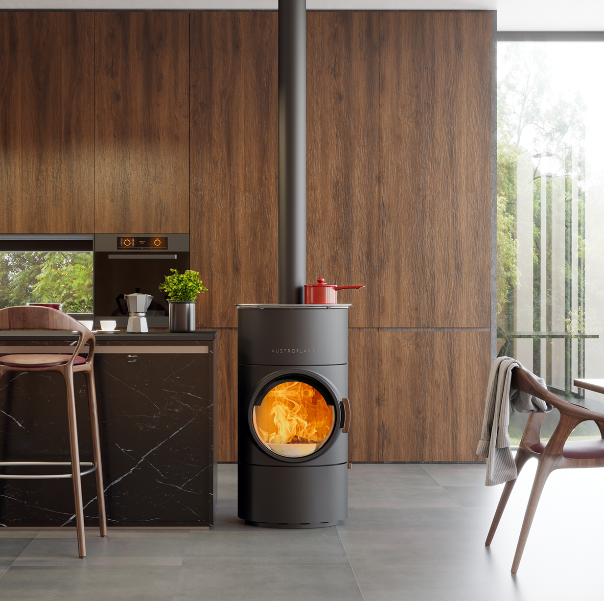 A modern dark gray cylindrical wood-burning stove with a glowing fire inside, set in a contemporary kitchen with wood-paneled walls and a tall metal chimney pipe extending upward.