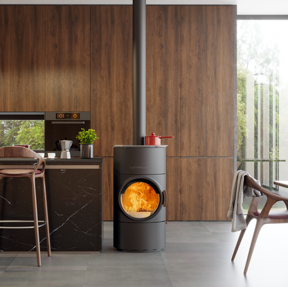 A modern dark gray cylindrical wood-burning stove with a glowing fire inside, set in a contemporary kitchen with wood-paneled walls and a tall metal chimney pipe extending upward.