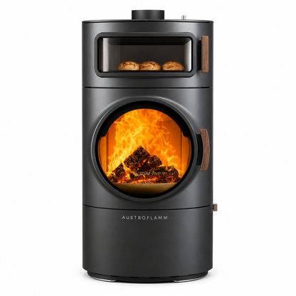 A black Austroflamm wood-burning stove with a glass door showing a glowing fire, and a smaller upper window with three baked pastries visible inside. The stove is photographed on a clean white background.