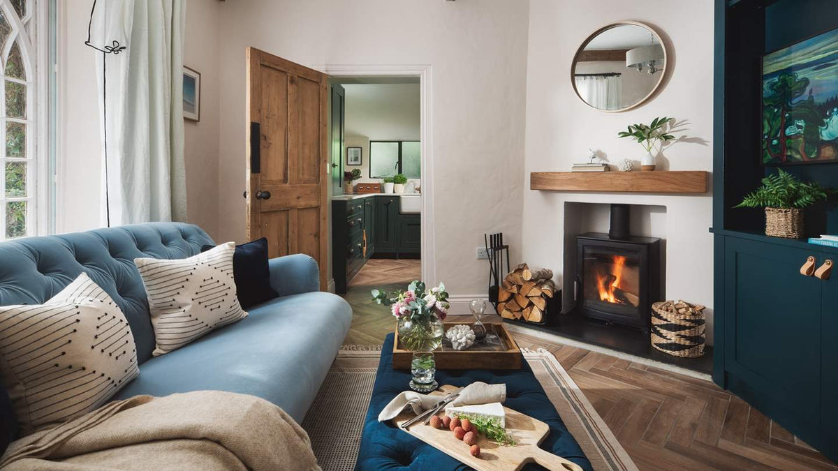 A beautifully styled living room featuring a tufted blue sofa with patterned throw pillows, a dark teal cabinet, a black wood-burning fireplace with a visible fire, and a wooden mantel.