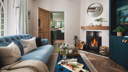 A beautifully styled living room featuring a tufted blue sofa with patterned throw pillows, a dark teal cabinet, a black wood-burning fireplace with a visible fire, and a wooden mantel.