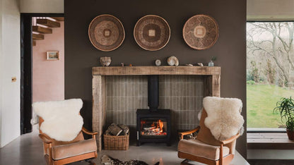 A cozy living room scene with a wood-burning fireplace centered against a dark wall, two upholstered armchairs with white sheepskin throws, and three woven circular wall decorations above the mantel. A window on the right shows a grassy outdoor area.
