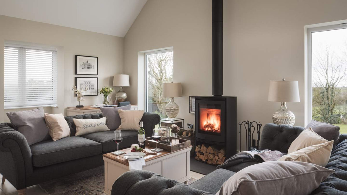 A modern black wood-burning stove with an open fire, set in a cozy living room with gray sofas, neutral-toned walls, and large windows.