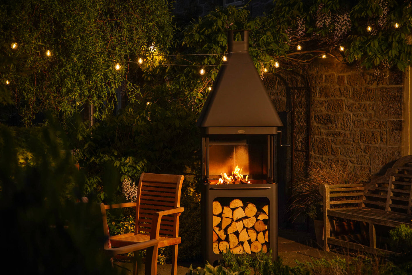 Carron Bellmount BBQ Fireplace Garden setting with a lit outdoor fireplace surrounded by chairs and a stone wall.