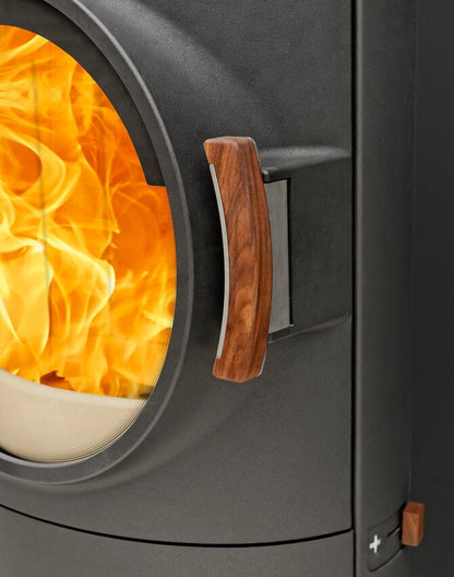 Close-up of a dark gray wood-burning stove with a circular glass door showing bright orange flames, a curved wooden handle, and part of the stove body on a dark background.
