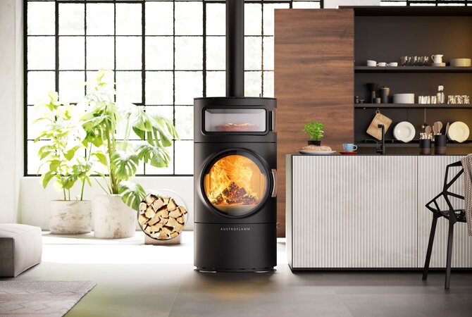 A modern black wood-burning stove with a large circular glass door showing a glowing fire, positioned in a contemporary kitchen and living space with plants, a wood cabinet, and a window.
