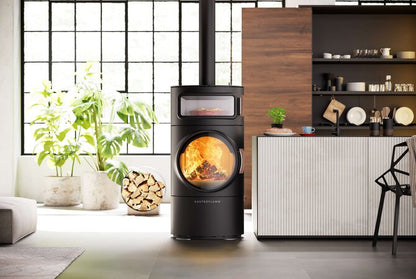 A modern black wood-burning stove with a large circular glass door showing a glowing fire, positioned in a contemporary kitchen and living space with plants, a wood cabinet, and a window.