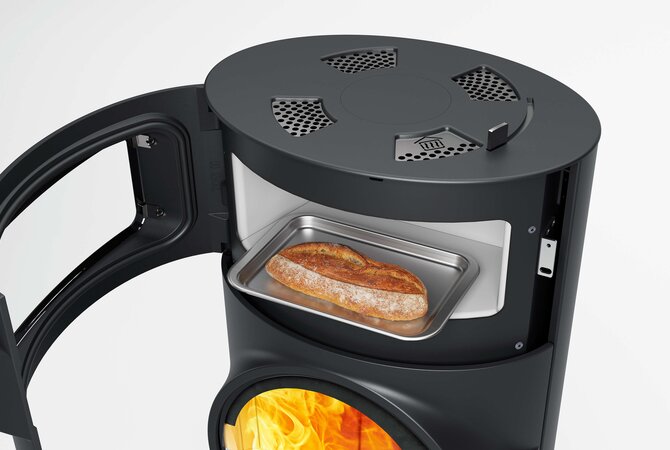 A black cylindrical wood-burning stove with its door open, showing a metal tray holding a loaf of bread inside the combustion chamber. The lower section reveals glowing orange flames.