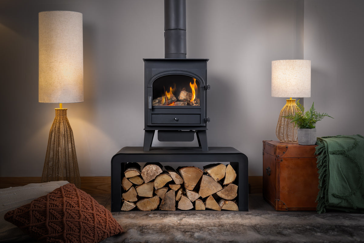 A black gas stove with a visible fire, set on a matching black firebox base filled with stacked logs. The scene is styled in a cozy room with two lit table lamps, a brown leather trunk, a green throw blanket, and a textured rug.