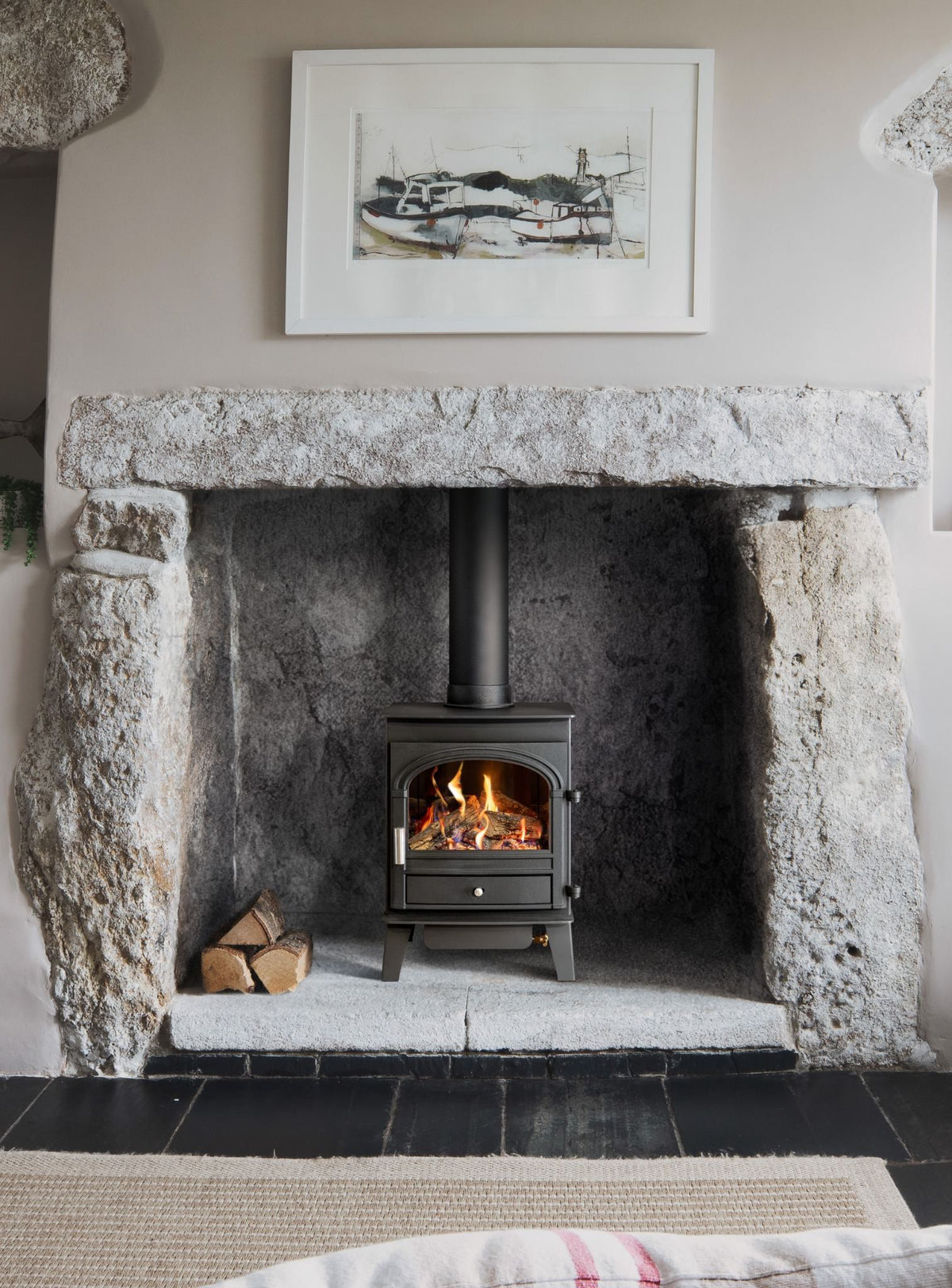 A black gas stove with a visible fire, set within a rustic stone fireplace. The scene includes a white-framed picture on the wall above, a stack of firewood, and a woven rug in the foreground.