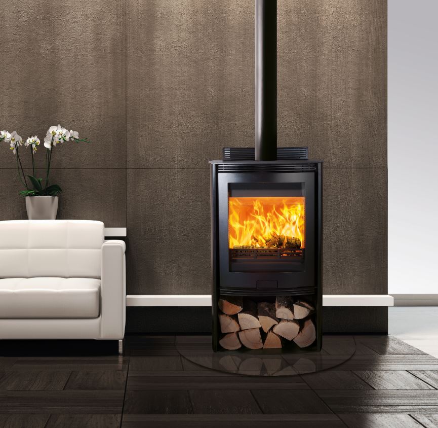 A modern black wood-burning stove with an open fire visible through the front glass door, set against a textured brown wall in a styled interior with a white sofa and dark wood flooring.