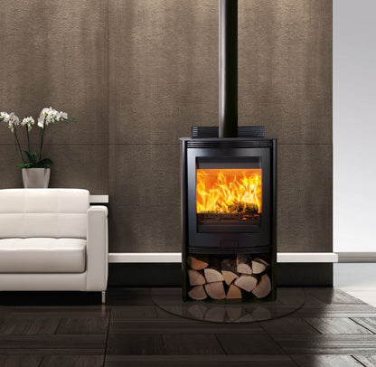 A modern black wood-burning stove with an open fire visible through the front glass door, set against a textured brown wall in a styled interior with a white sofa and dark wood flooring.