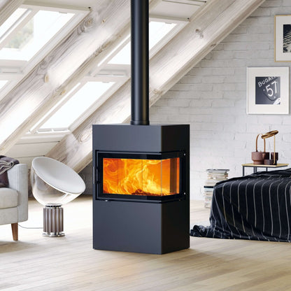 A modern black wood-burning stove with a glass front door showing a bright fire, set in a bright, light-filled attic bedroom with white brick walls and large skylights.