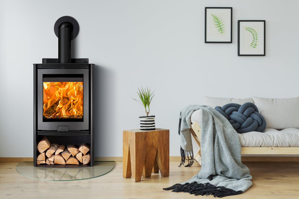 A black wood-burning stove with a visible fire, set in a modern living room next to a wooden stool, a gray throw blanket, and a sofa.