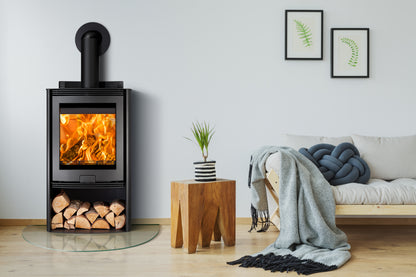 A black wood-burning stove with a visible fire, set in a modern living room next to a wooden stool, a gray throw blanket, and a sofa.