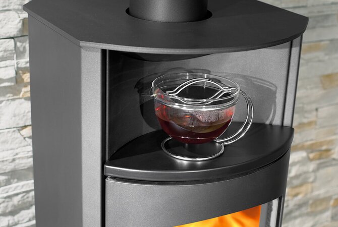 A modern black wood-burning stove with a glass viewing window, shown with a glass teacup on its shelf against a light stone wall background.