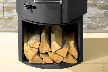 A black outdoor fire pit stove with a wood-burning compartment filled with cut firewood, set against a light stone wall and tiled floor.