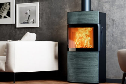 A modern wood-burning stove with a dark textured finish and a glowing orange fire visible through the glass door, set in a contemporary living room next to a white armchair.