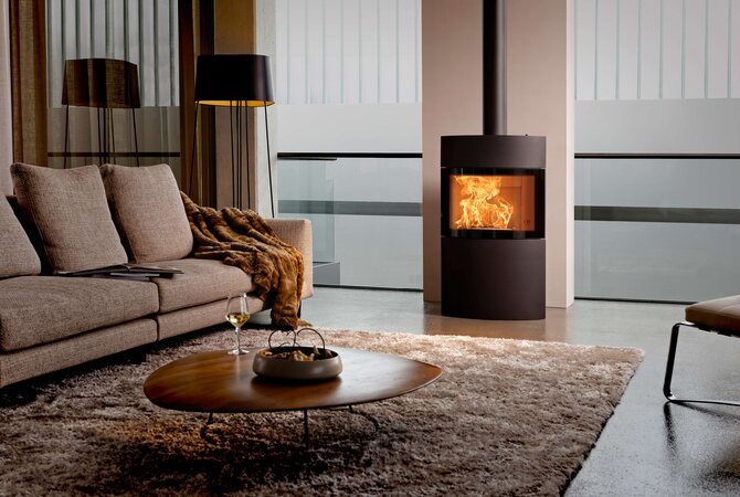 A modern cylindrical fireplace with a visible orange fire inside, set in a contemporary living room next to a beige sofa and a round wooden coffee table.