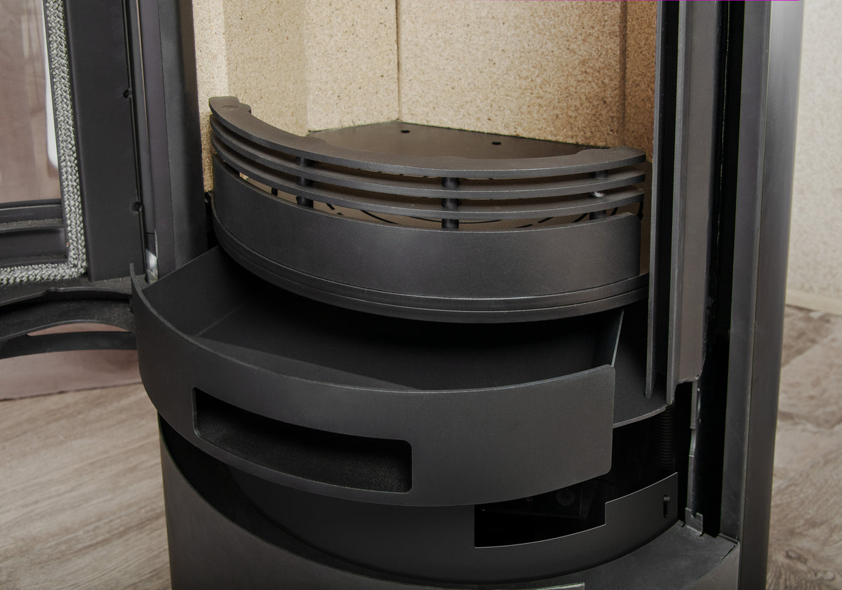 Close-up of a black metal circular fireplace insert or burner assembly inside a stove or heater chamber, with beige textured walls and a wood-look floor visible.