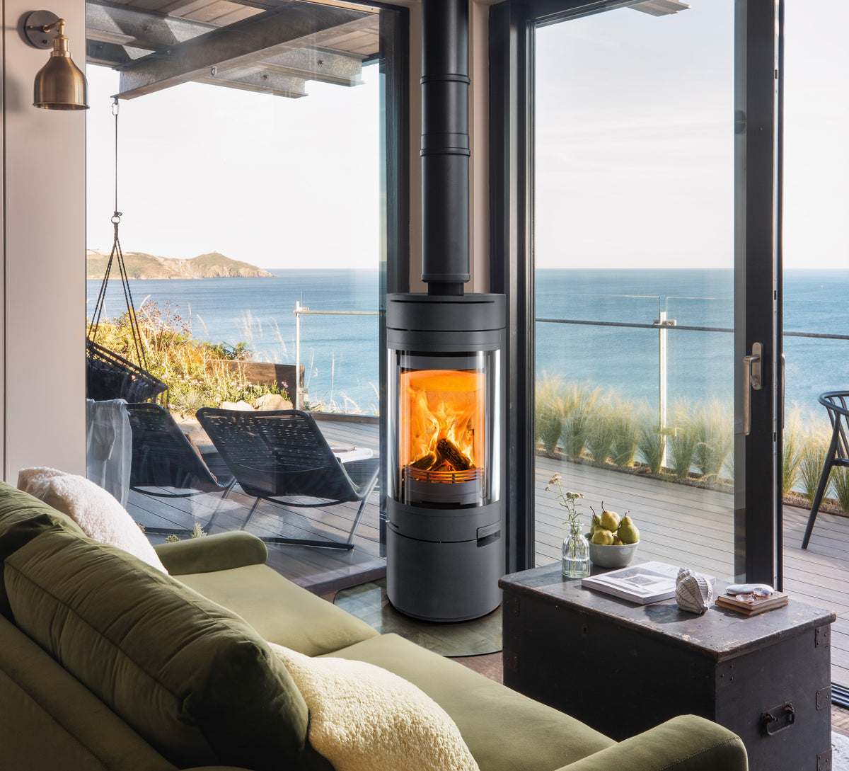 A modern black wood-burning stove with a glass front and glowing orange fire, positioned in a stylish living room with a green sofa and large windows overlooking the ocean.