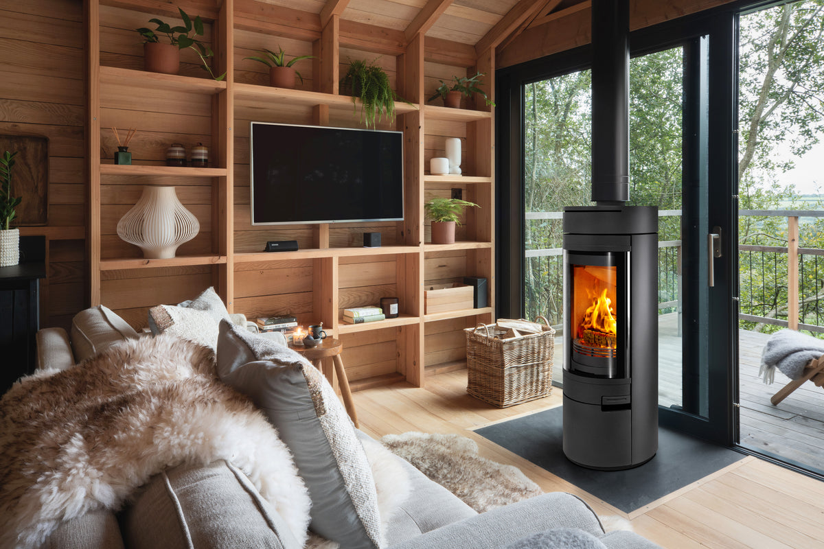 A modern black wood-burning stove with a glowing fire, set in a cozy, contemporary cabin living room with wooden walls, built-in shelves, and large glass doors looking out to a green forest.