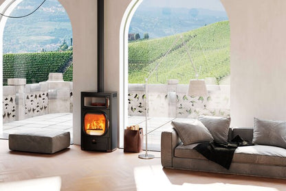 A black wood-burning stove with a glowing fire, set in a modern living room with a gray sofa and large arched windows overlooking a vineyard landscape.