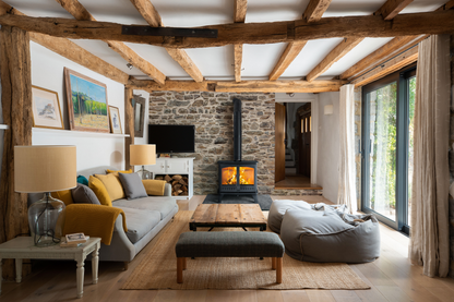A cozy, rustic living room featuring a gray sofa with mustard yellow and gray pillows, a wooden coffee table, a gray beanbag chair, a black wood-burning stove, and a stone wall in the background. The room has exposed wooden ceiling beams and large windows with beige curtains.