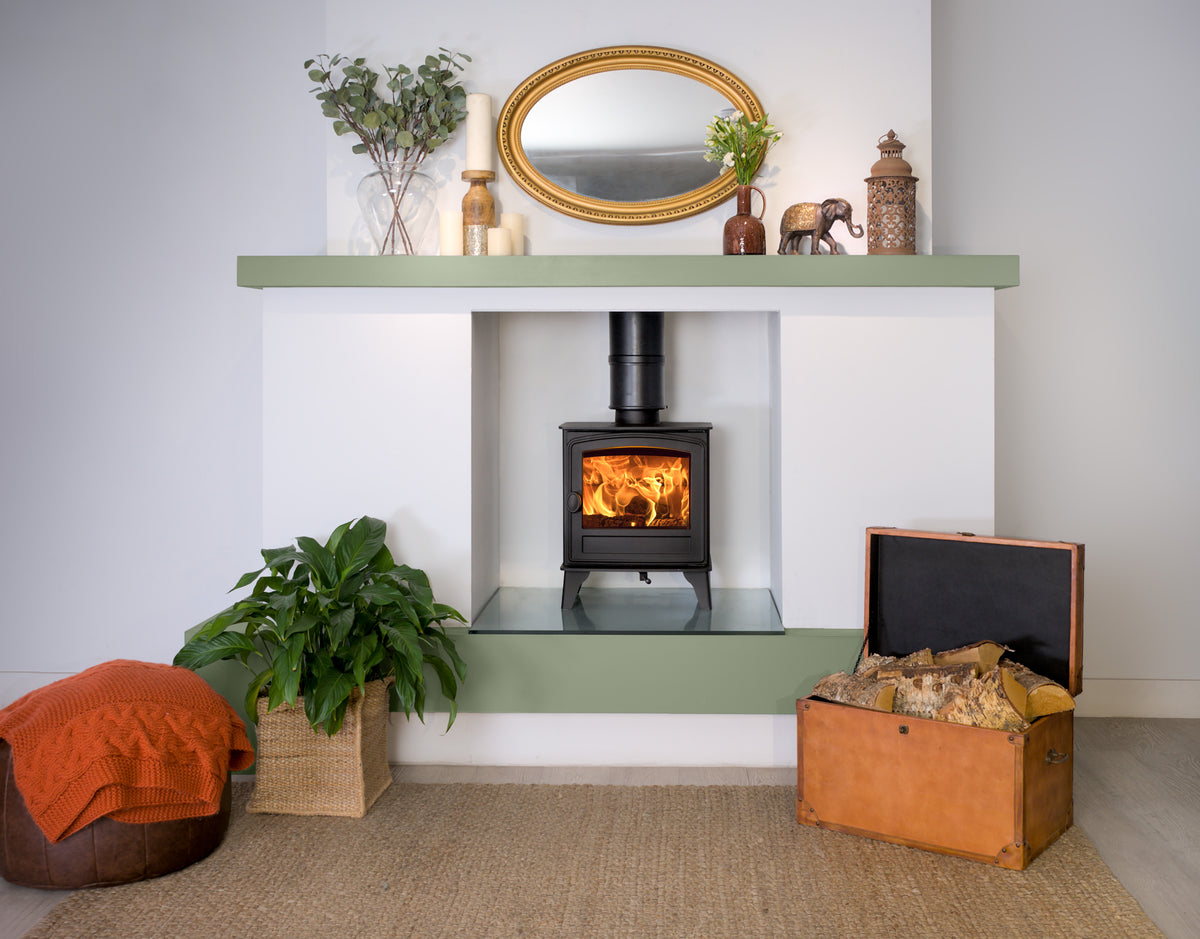 A modern wood-burning stove insert set into a white wall niche with sage green mantel and base shelves. The scene is styled as a cozy living room with a lit fire, a gold-framed mirror, decorative plants, a leather trunk, and an orange pouf on a woven rug.